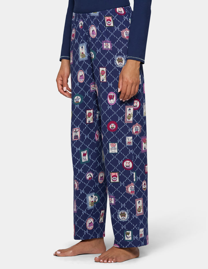 A person wearing navy blue pajama pants with a repeating vintage postcard and diamond lattice pattern, paired with a navy long-sleeve top, shown against a light gray studio background.