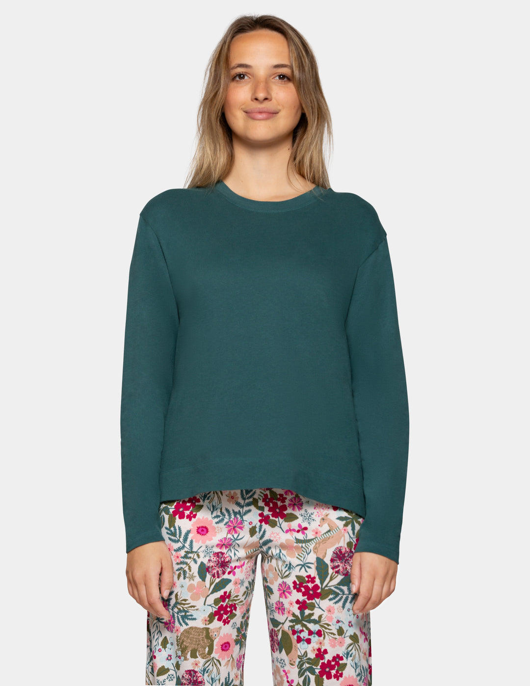 A woman modeling a dark teal green long-sleeve crewneck top, paired with pink and white floral print pants, against a solid light gray background.