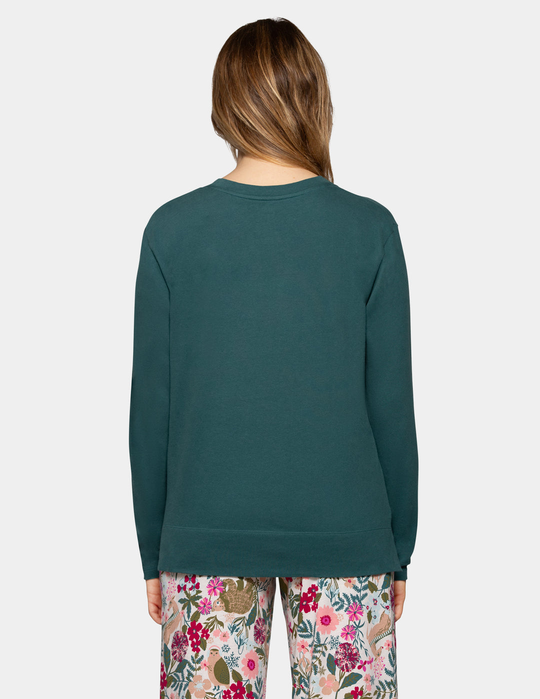 A woman shown from the back wearing a dark teal long-sleeve crewneck sweatshirt and floral pajama pants against a light gray studio background.