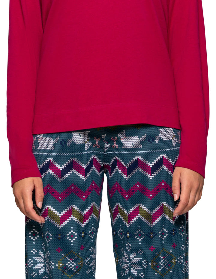 A person wearing a bright pink long-sleeve top and patterned knit pants. The pants have a dark teal base with a multicolor holiday-style geometric and animal print in pink, white, navy, and olive green.