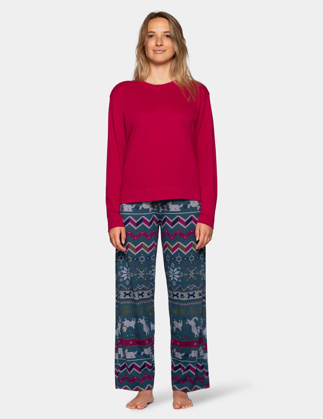 A woman standing against a light gray studio background wearing a bright pink long-sleeve top and patterned pajama pants with a blue, gray, and magenta Fair Isle design featuring snowflakes, geometric stripes, and animal motifs.