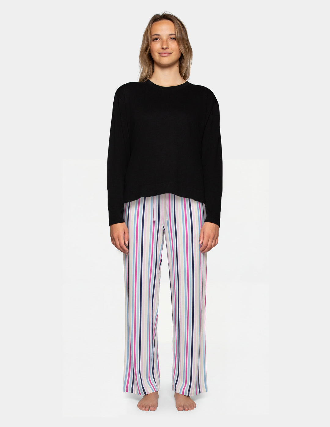A woman stands against a light gray studio background wearing a black long-sleeve top and white pants with vertical pink, purple, and blue stripes.