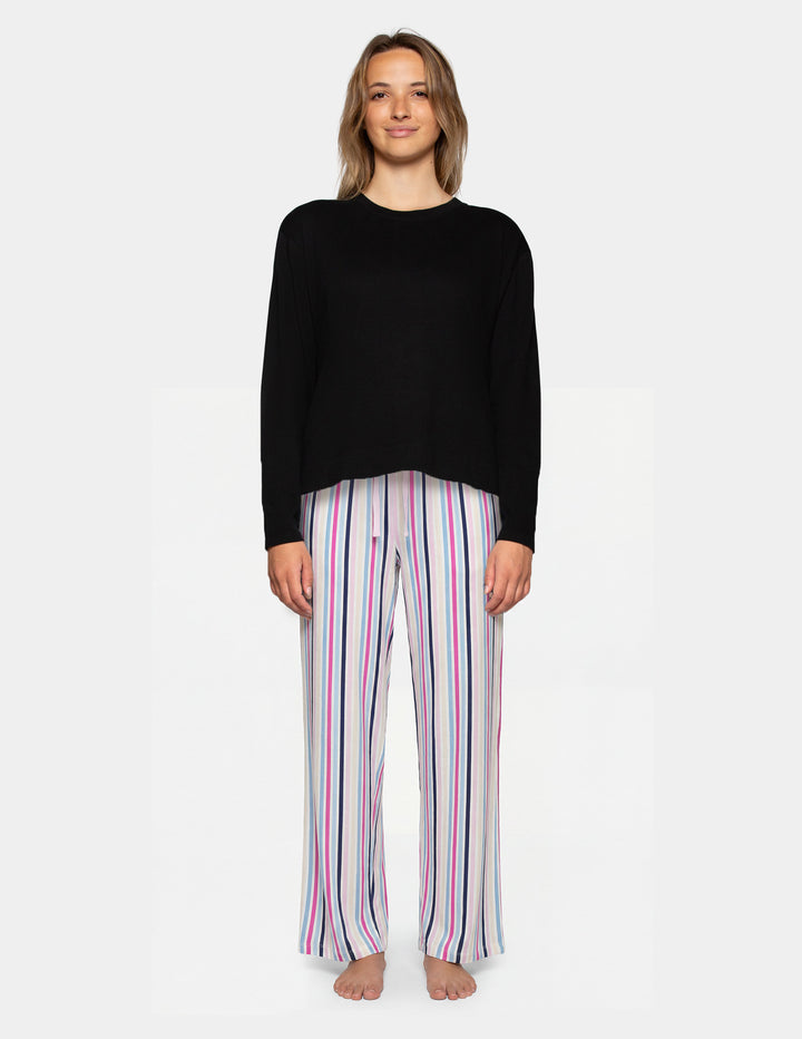 A woman stands against a light gray studio background wearing a black long-sleeve top and white pants with vertical pink, purple, and blue stripes.