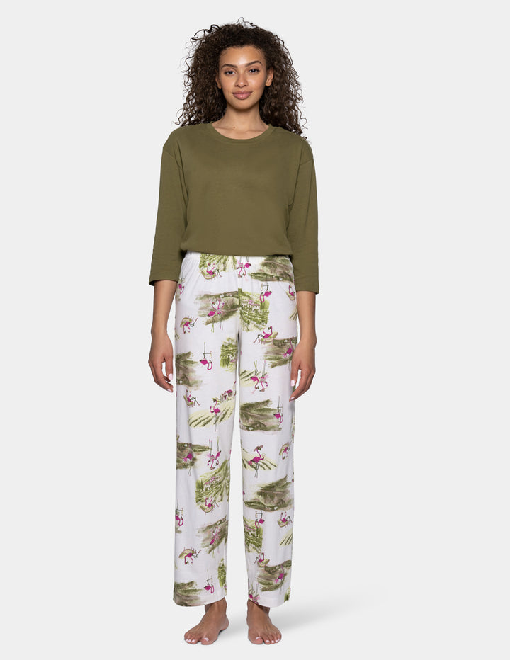 A woman wearing an olive green pajama top and white pj pants with an allover print of green palm leaves and pink flamingos, shown from the waist down against a light background.