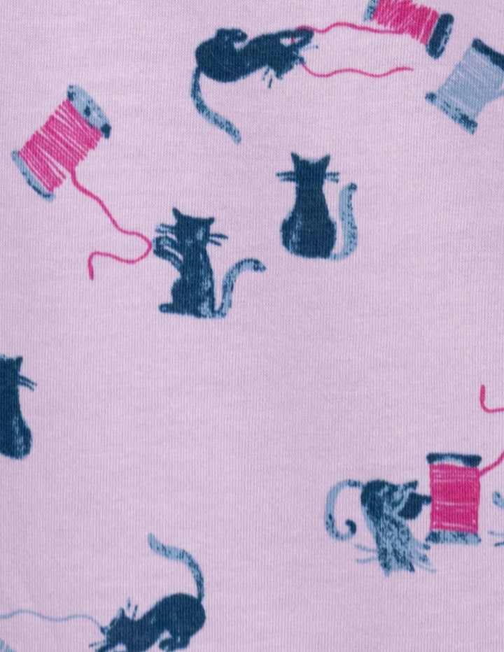 A close-up of a light purple fabric with a repeating pattern of cats playing with pink and blue spools of thread.