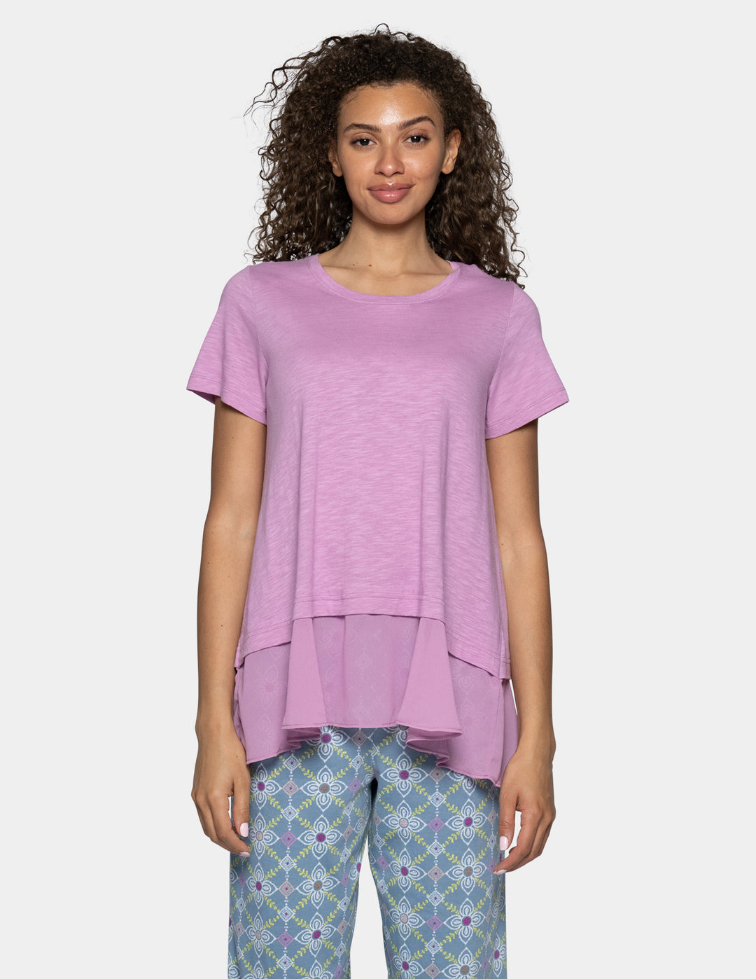 A woman modeling a short-sleeve, high-low tiered top in a dusty lavender purple, paired with patterned blue and purple pants, against a light gray background.