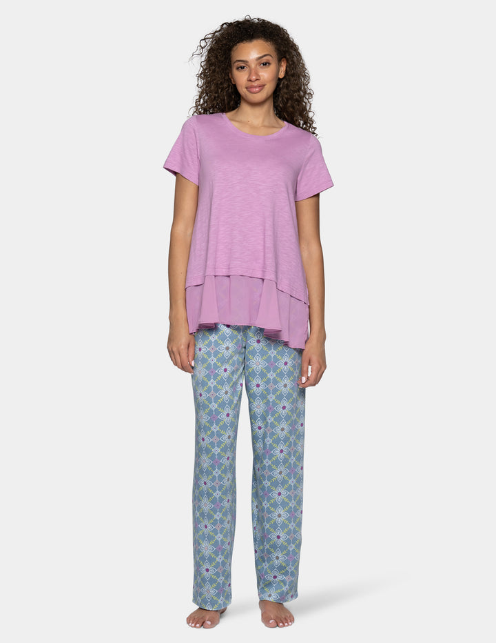 A woman modeling a short-sleeve, high-low tiered top in a dusty lavender purple, paired with patterned blue and purple pants, against a light gray background.