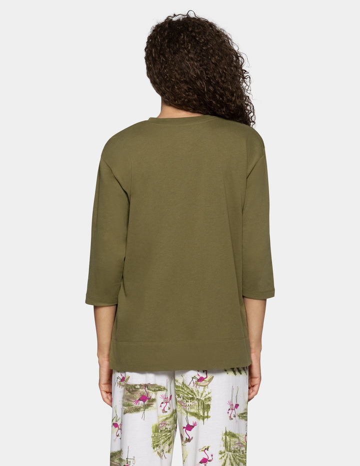 A woman shown from the back wearing an olive green three-quarter sleeve top and white tropical print pants against a light gray background.