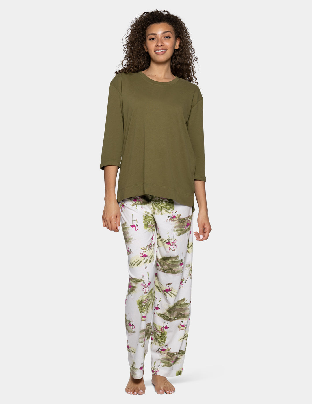 A woman modeling a two-piece pajama set consisting of an olive green long-sleeve top and white pants with a green tropical leaf and pink flamingo print, against a light gray background.