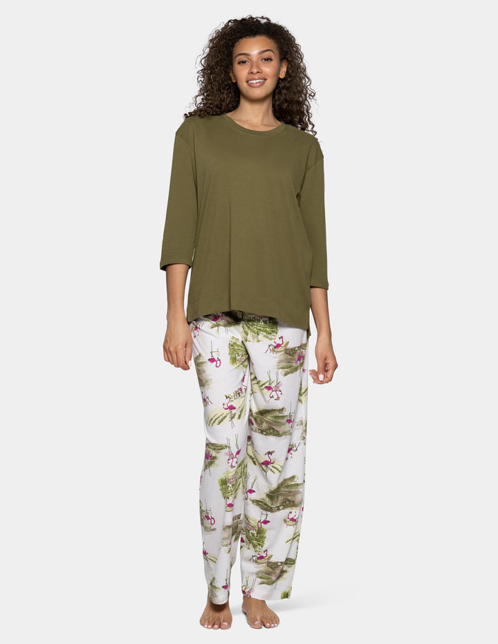 A woman modeling a two-piece pajama set consisting of an olive green long-sleeve top and white pants with a green tropical leaf and pink flamingo print, against a light gray background.