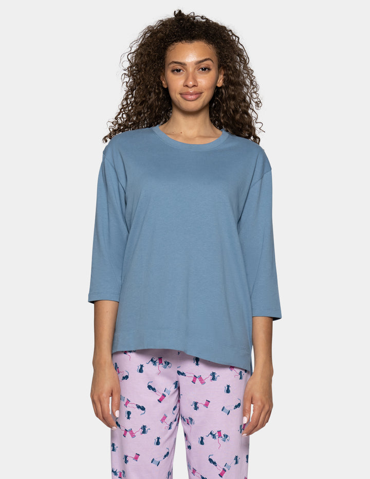 A woman modeling a dusty blue three-quarter sleeve t-shirt and pink patterned pajama pants against a light gray background.