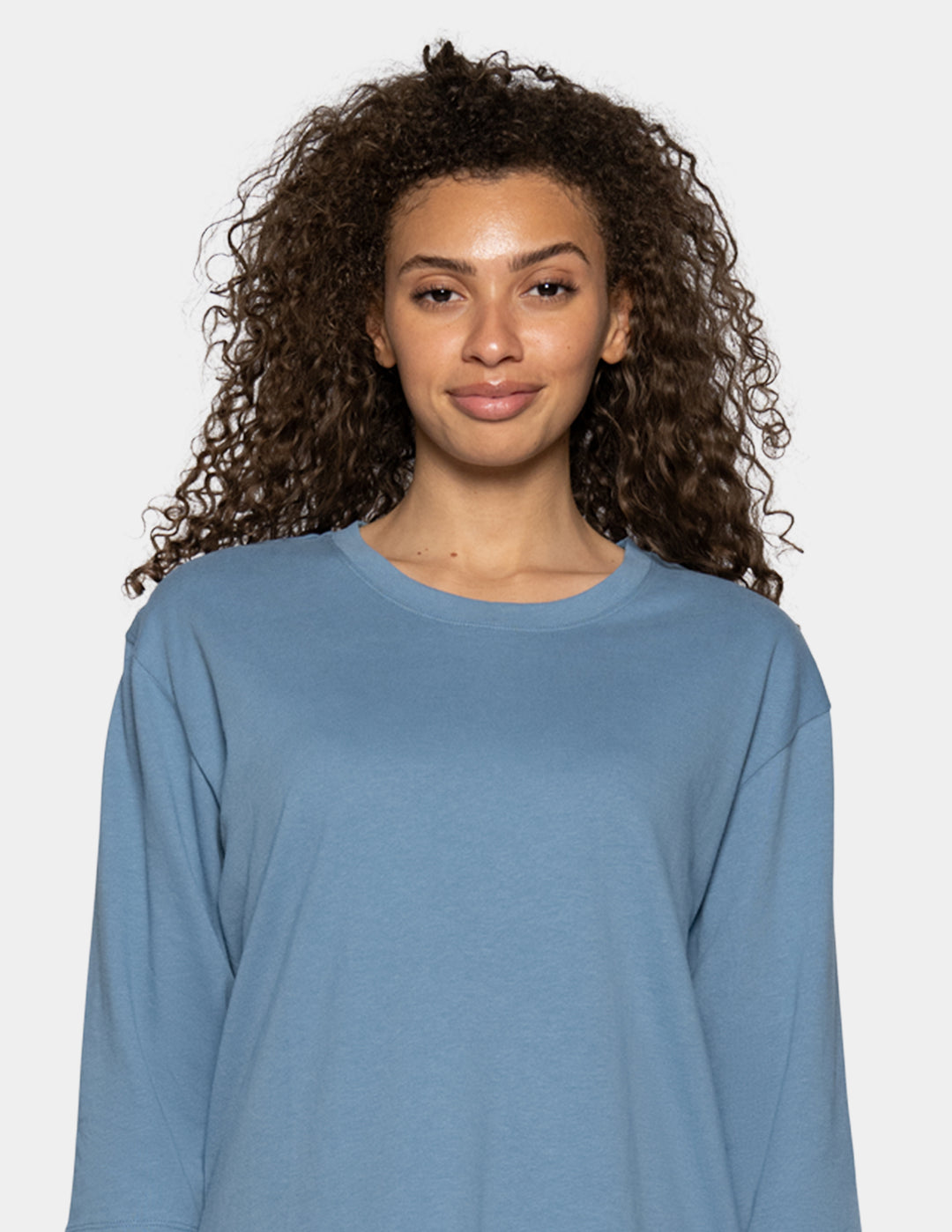 A woman wearing a dusty blue long-sleeve crewneck top, photographed against a solid light gray background.