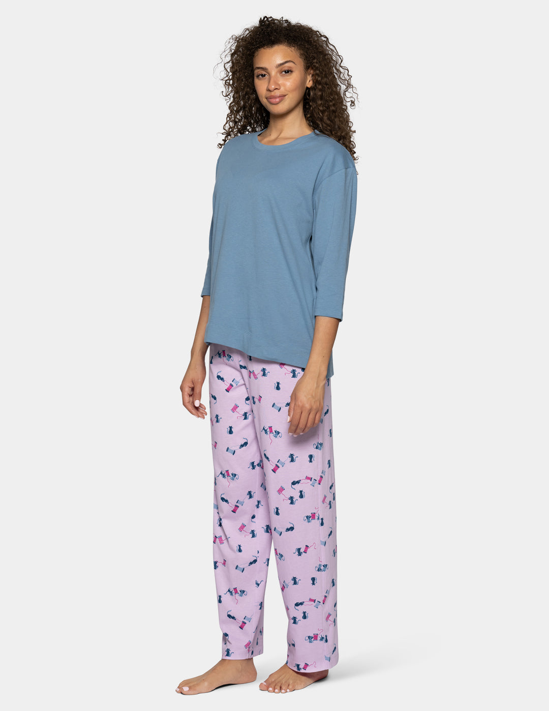 A woman modeling a two-piece pajama set consisting of a dusty blue long-sleeve top and light pink pants with a small multicolor pattern, shot against a light gray studio background.
