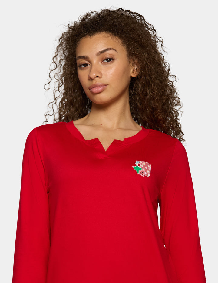 A woman with curly brown hair wearing a bright red long-sleeve top with a split V-neckline and a small colorful embroidered chest detail, photographed against a light gray studio background.