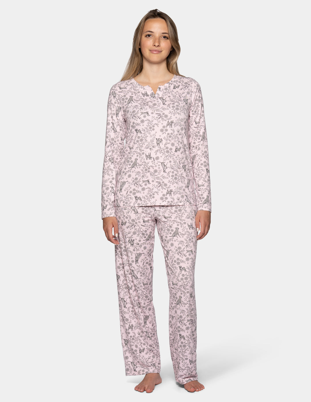 A woman wearing a light pink long sleeve pajama top featuring a floral and dog pattern, with matching full length pajama pants against a gray background.