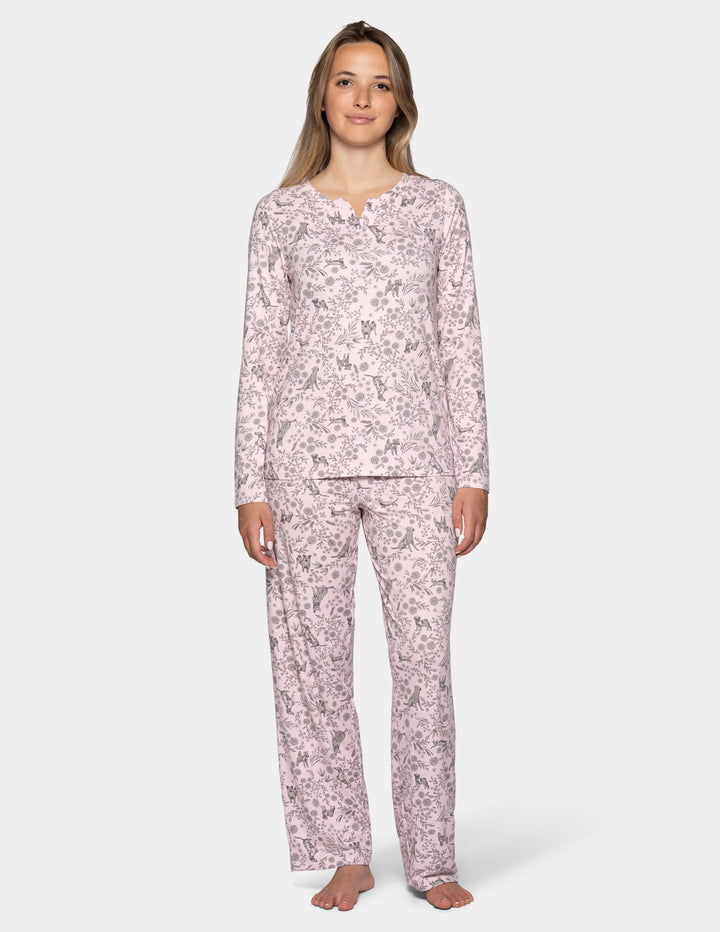 A woman wearing a light pink long sleeve pajama top featuring a floral and dog pattern, with matching full length pajama pants against a gray background.