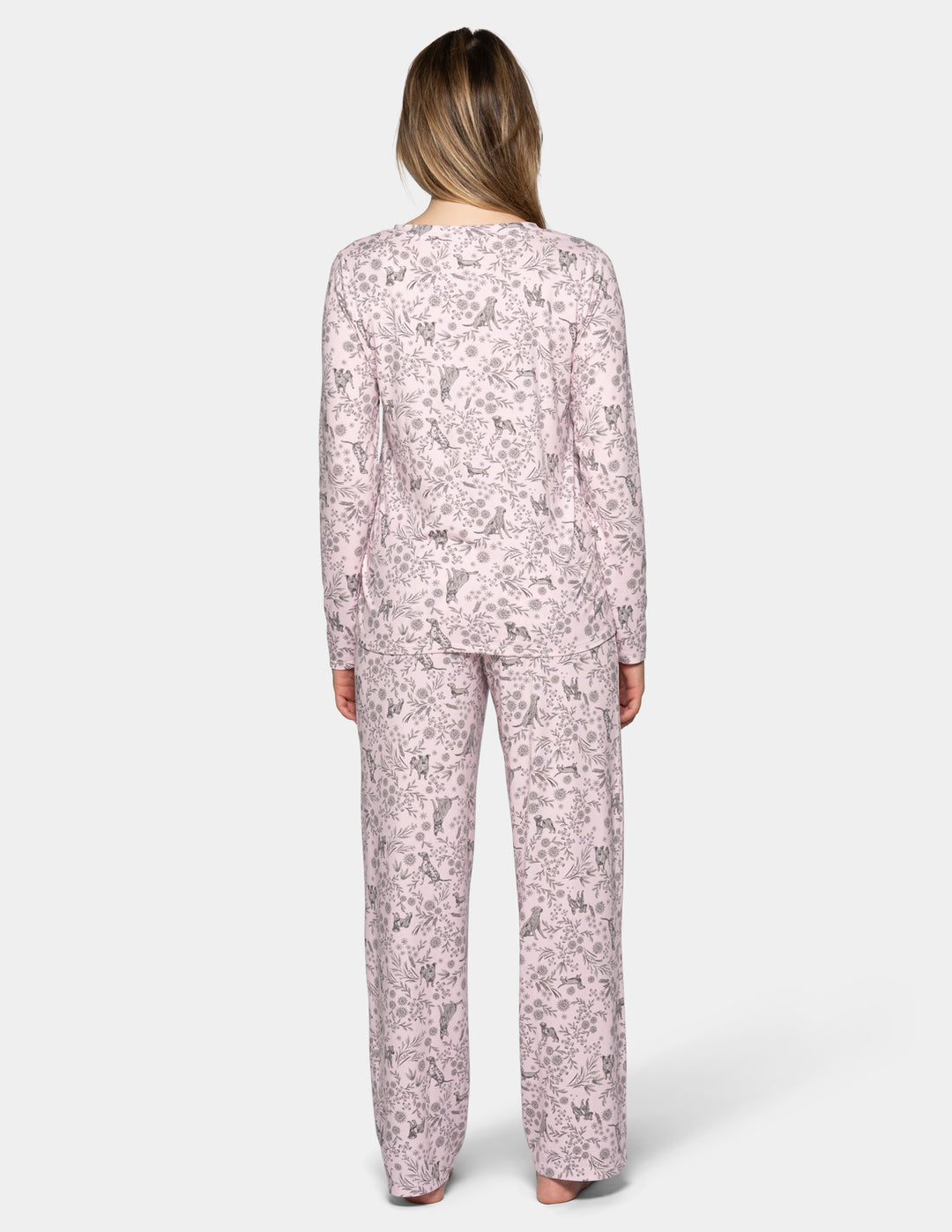 A backside view of a woman wearing a light pink long sleeve pajama top featuring a floral and dog pattern, with matching full length pajama pants against a gray background.