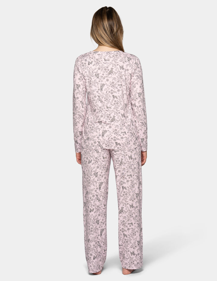 A backside view of a woman wearing a light pink long sleeve pajama top featuring a floral and dog pattern, with matching full length pajama pants against a gray background.