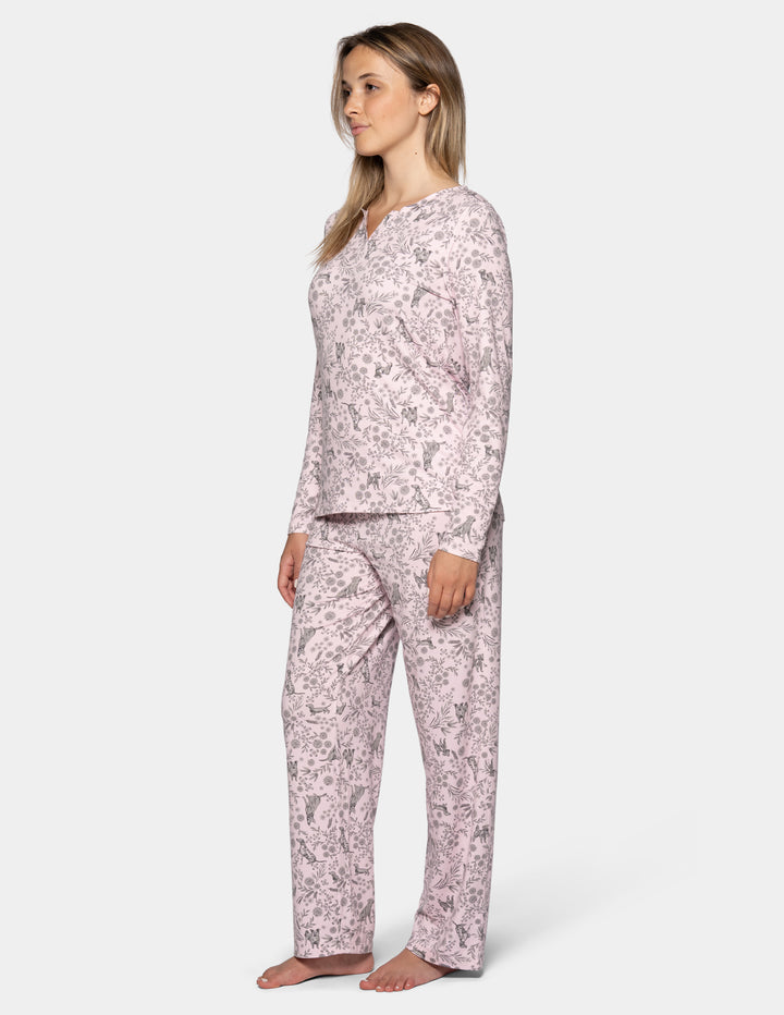 A sideview of a woman wearing a light pink long sleeve pajama top featuring a floral and dog pattern, with matching full length pajama pants against a gray background.