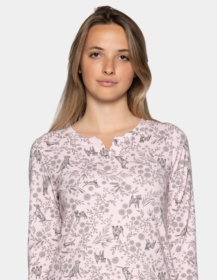 A woman wearing a light pink long sleeve pajama top featuring a floral and dog pattern against a gray background.
