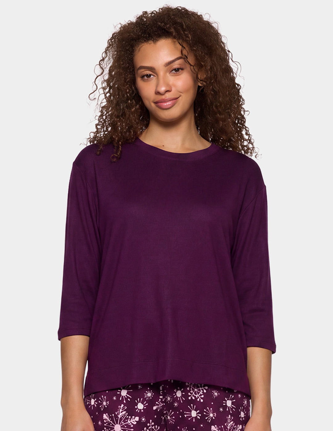 A woman with curly brown hair wearing a dark purple, three-quarter sleeve top and matching purple snowflake pants, photographed against a solid light gray background.