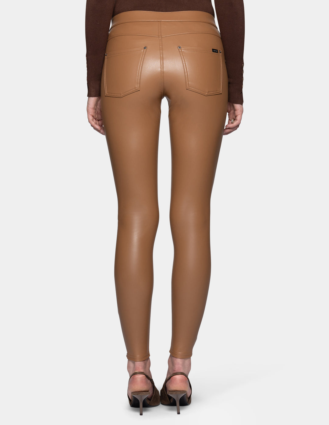 A backside view of a model wearing tan faux leather leggings and a long sleeve brown top, paired with suede brown heels against a light grey background.