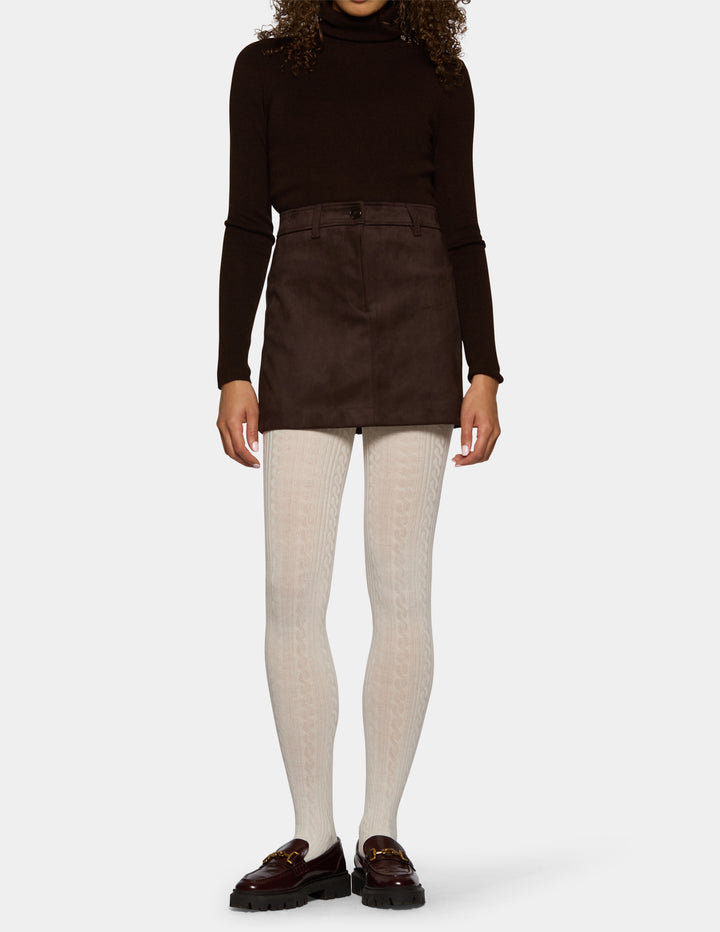 A model wearing ivory textured tights, a brown skirt, and a long sleeve black top against a light gray studio background.
