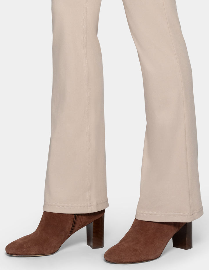 Close-up of a person's legs wearing beige bootcut pants and brown suede block-heel ankle boots against a light gray background.