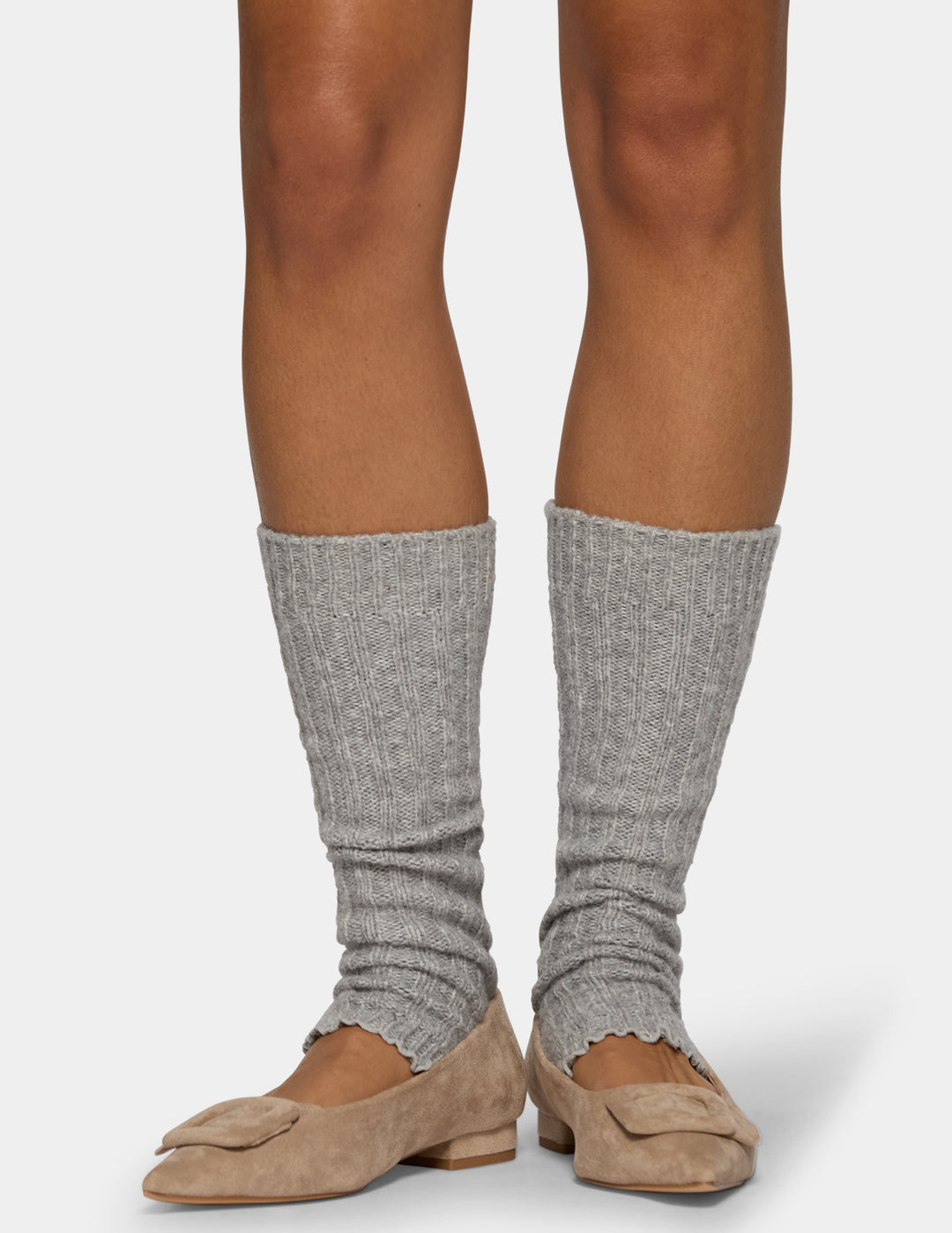 A model's legs wearing gray ribbed leg warmers with scalloped edges, paired with tan suede flat shoes, against a light gray studio background.