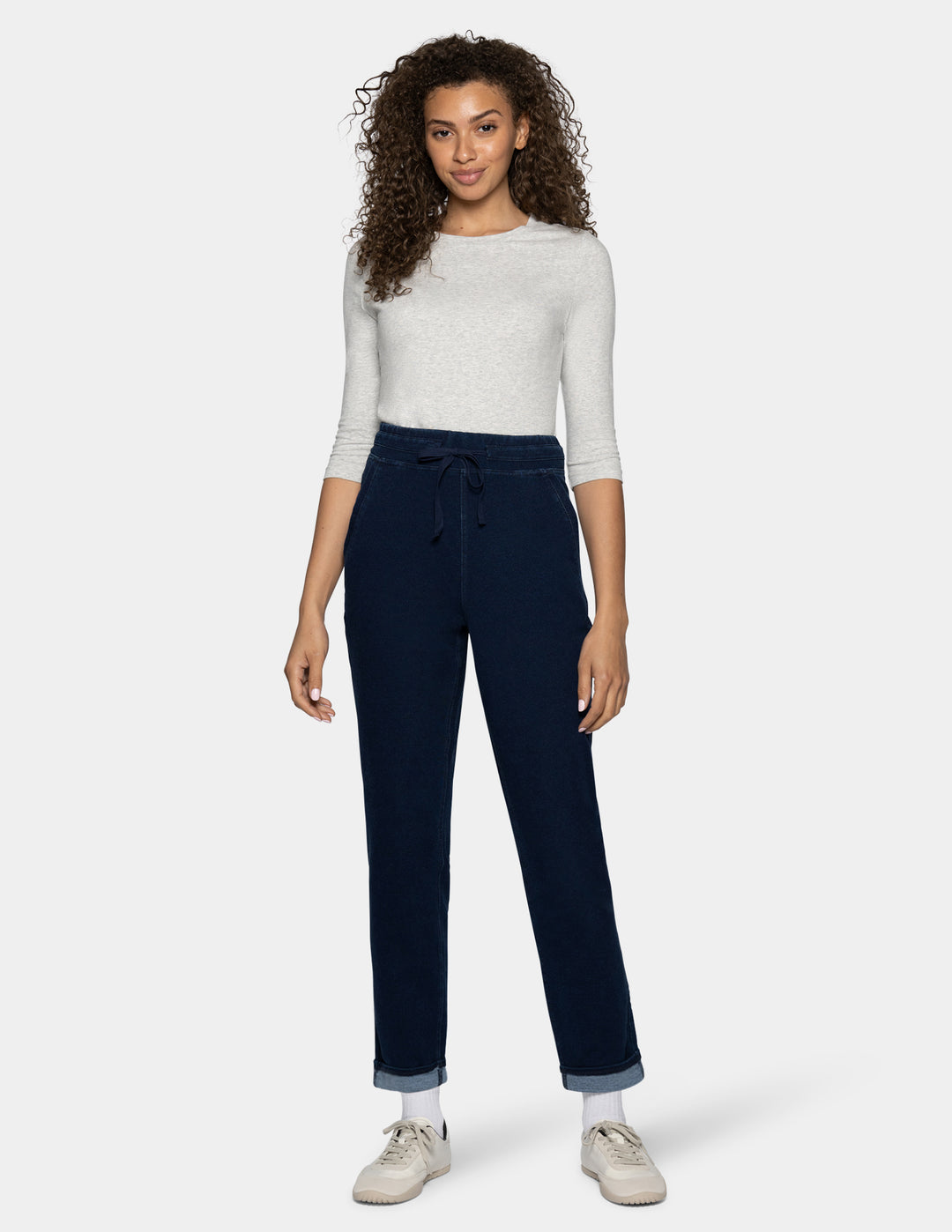 A woman modeling dark blue drawstring pants with a light gray knit top and white sneakers, standing against a light gray background.
