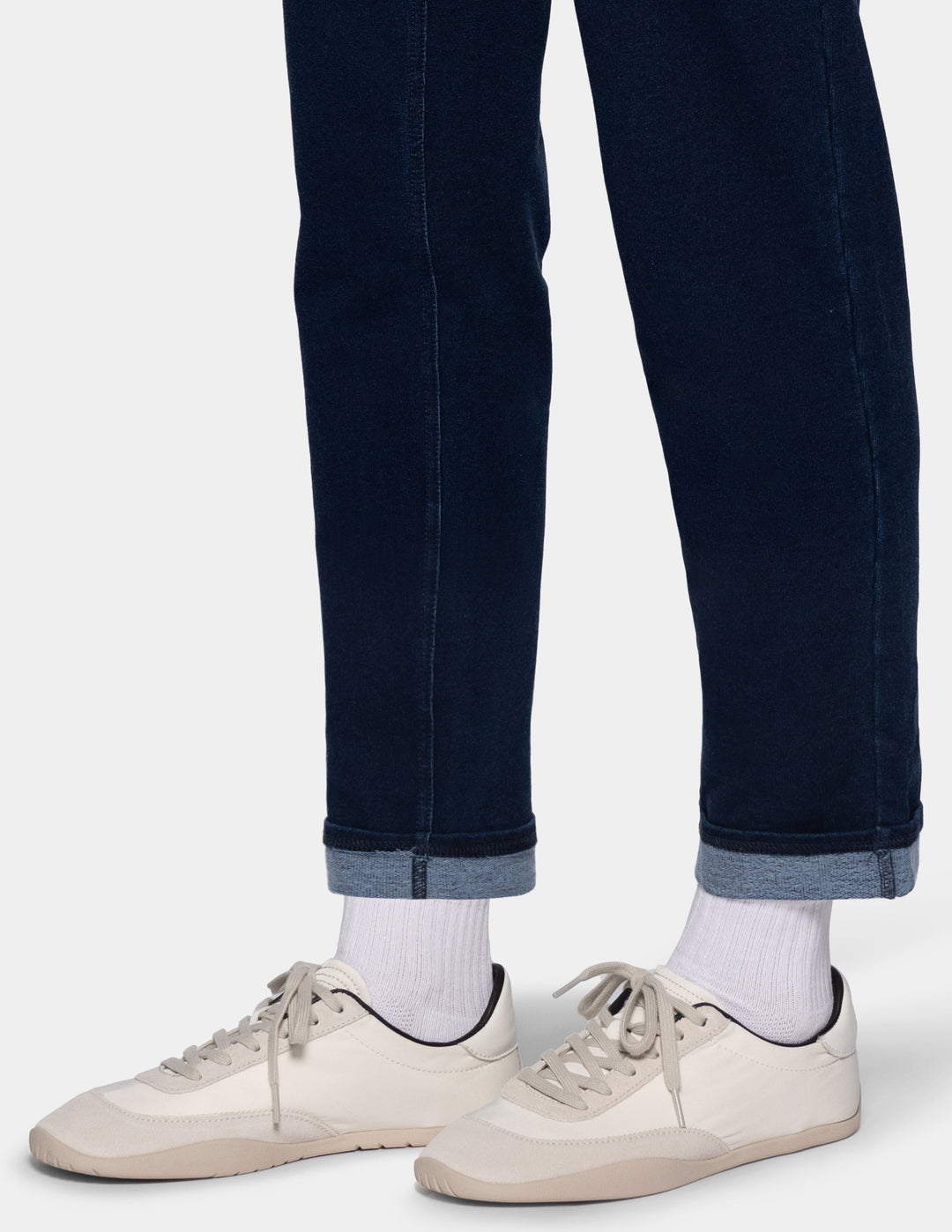 Close-up of a person's legs wearing dark blue joggers with a cuffed hem, white crew socks, and light beige low-top sneakers against a light gray background.
