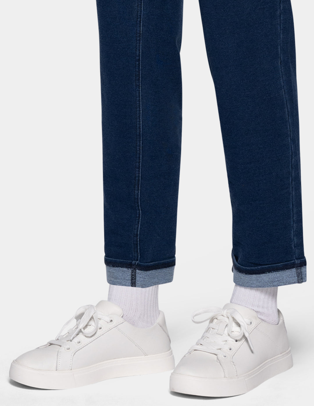 A close-up of a person's legs wearing dark blue cuffed joggers, white socks, and white low-top sneakers against a light gray background.