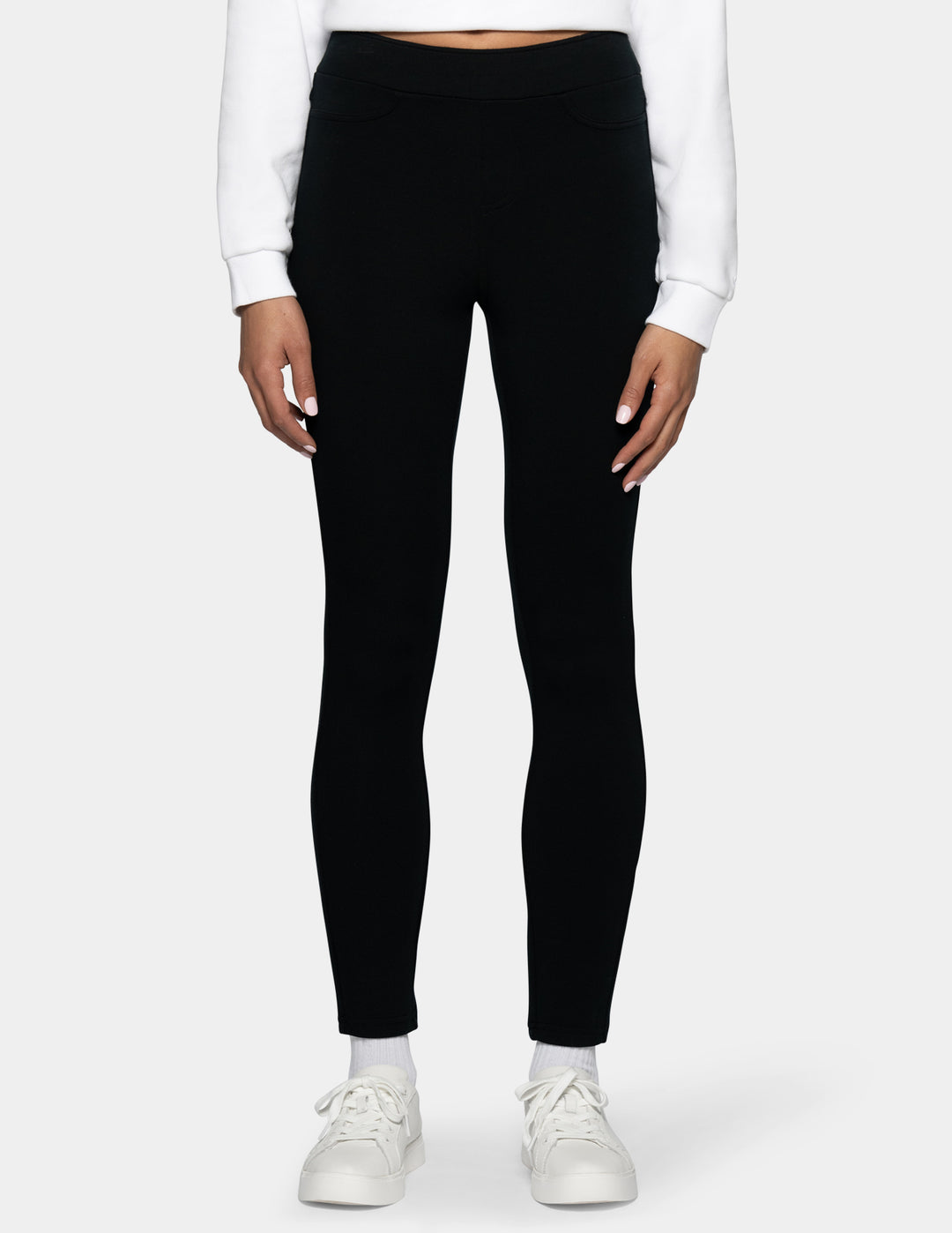 A model wearing black full-length leggings, styled with a white long-sleeve crop top and white sneakers, shown from the waist down against a light gray background.