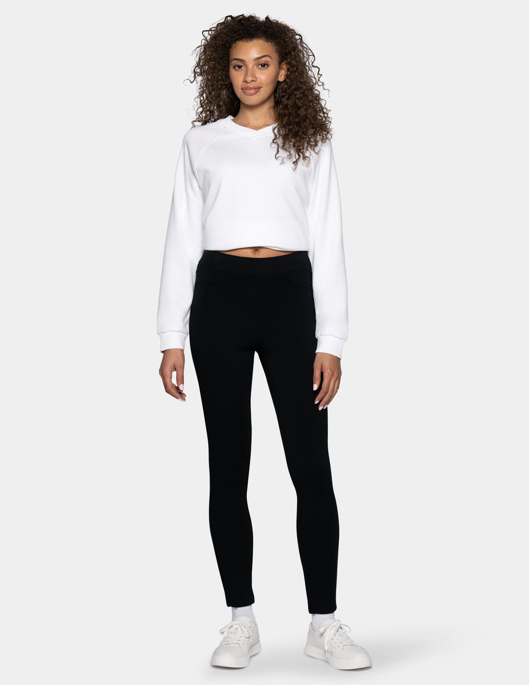 A woman modeling a white cropped long-sleeve sweatshirt and black leggings with white sneakers, standing against a light gray studio background.