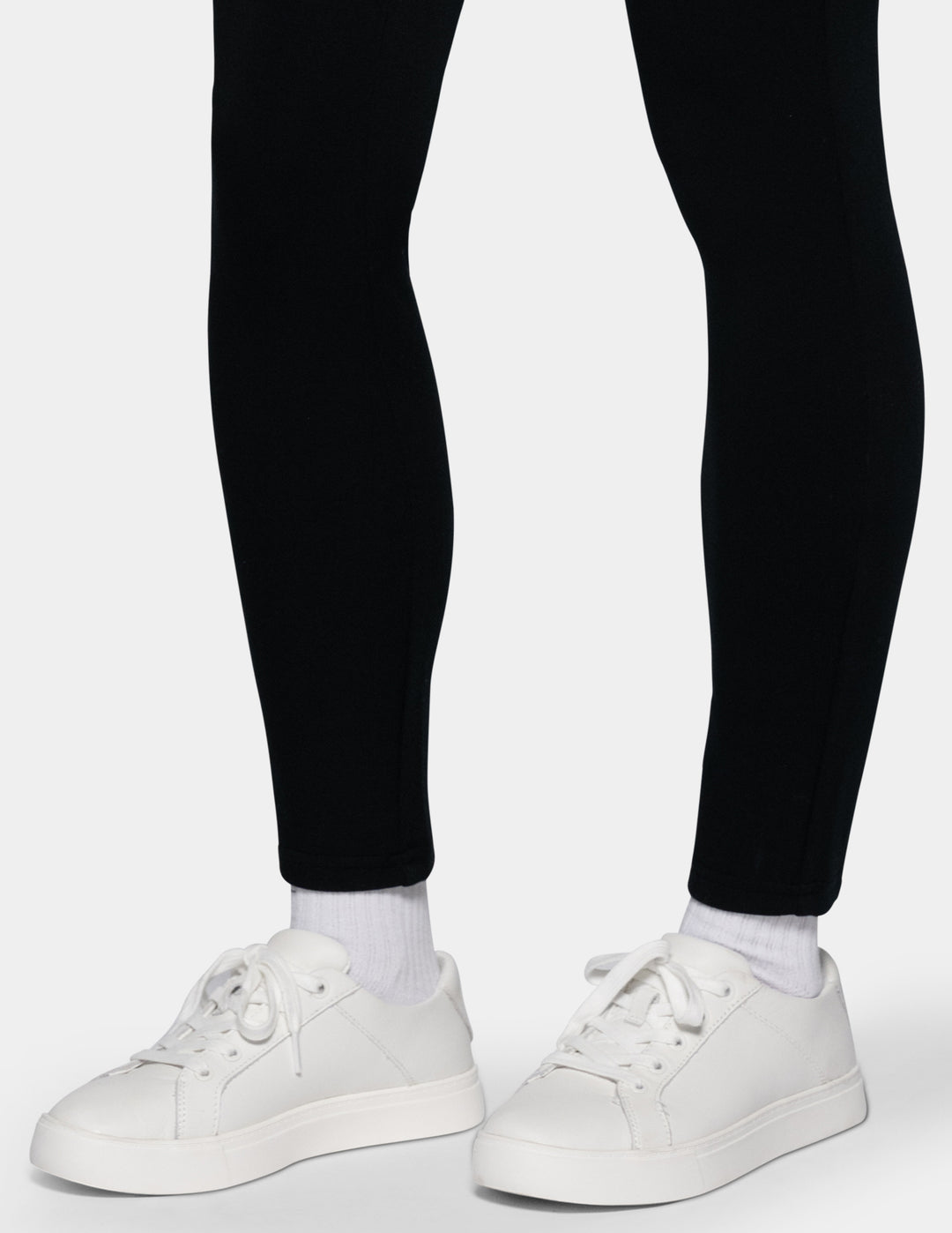 White low-top sneakers worn with white socks and black leggings, shown from the feet up against a light gray background.