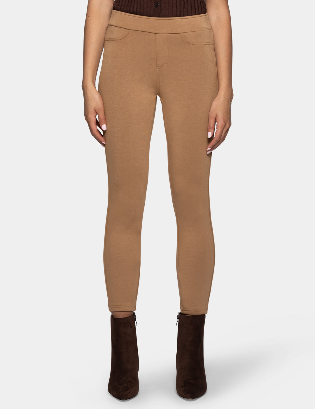 A model wearing slim-fit, ankle-length tan leggings and dark brown ankle boots, set against a light gray background.