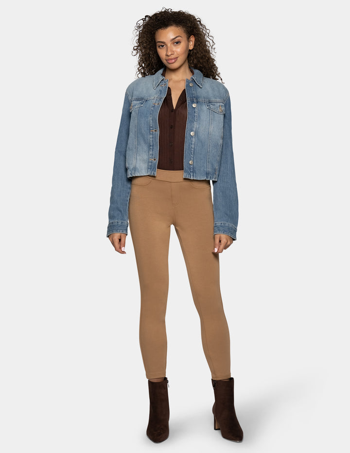 A model wearing a cropped light blue denim jacket over a dark brown top, paired with tan leggings and dark brown ankle boots, standing against a light gray background.