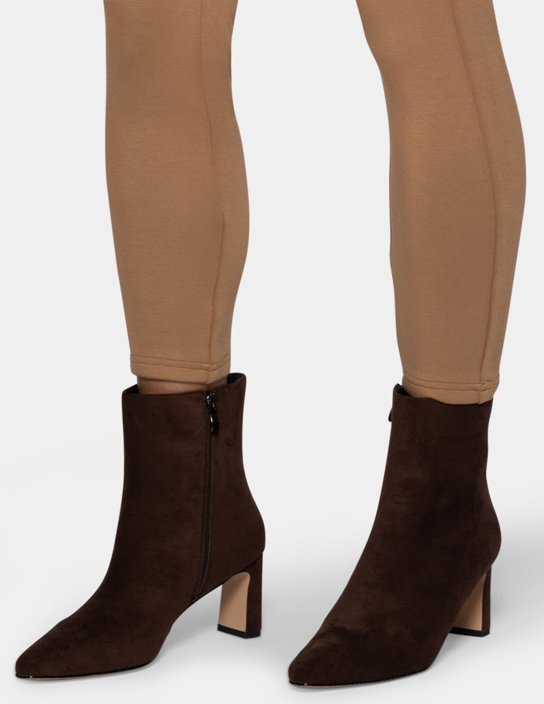 A pair of dark brown suede ankle boots with block heels, shown on a model wearing tan leggings against a light gray background.