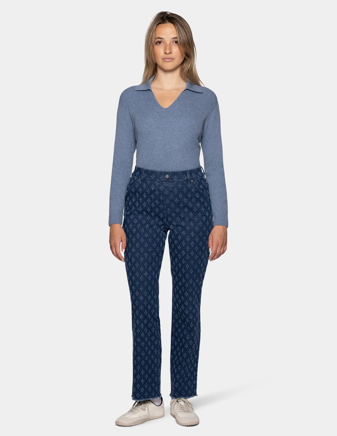 A woman stands against a light gray studio background wearing a heathered blue long-sleeve top, dark blue patterned pants, and off-white sneakers.