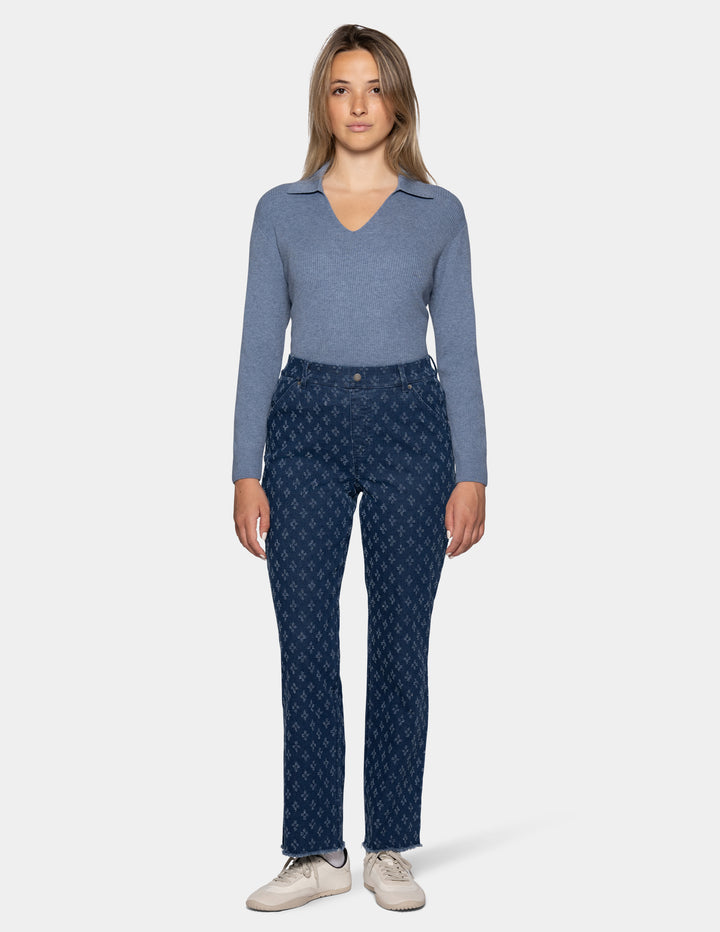 A woman stands against a light gray studio background wearing a heathered blue long-sleeve top, dark blue patterned pants, and off-white sneakers.
