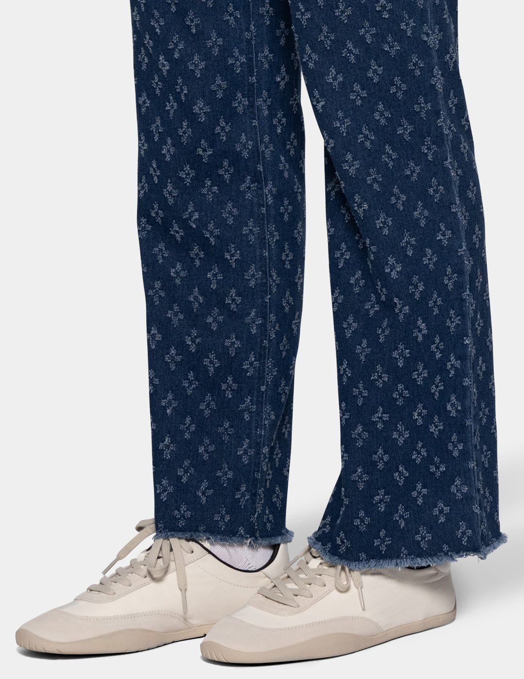 Close-up of a person's legs wearing dark blue textured patterned jeans with a raw frayed hem and cream-colored sneakers, shown against a light gray studio background.