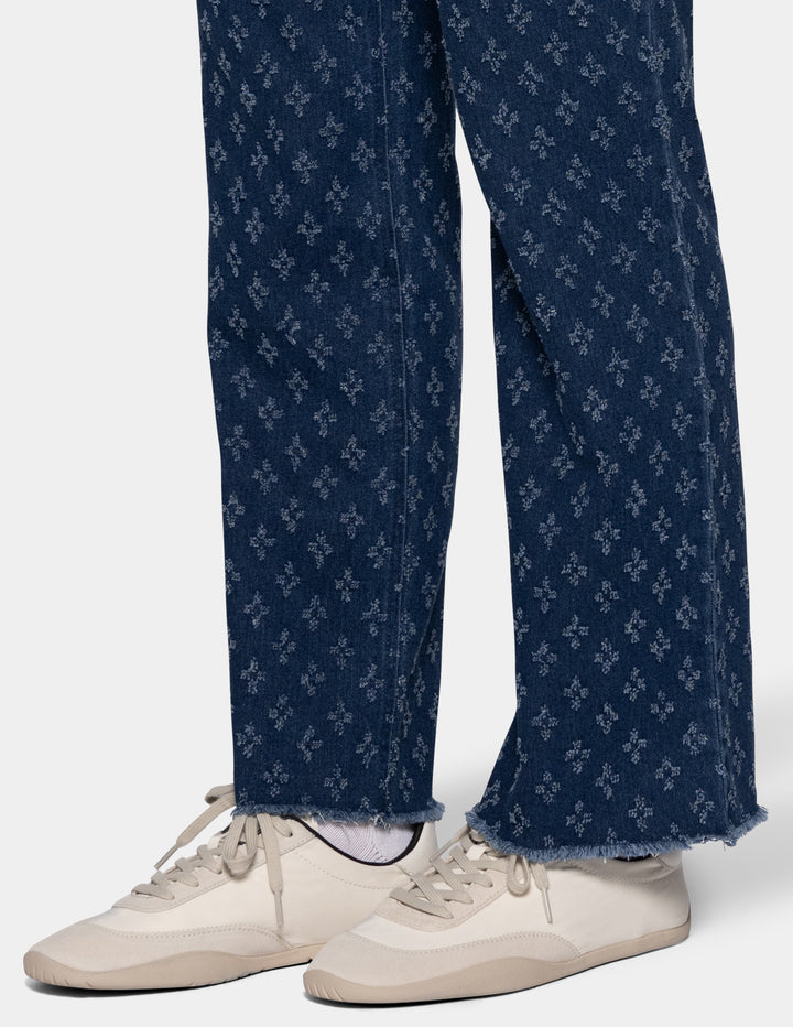 Close-up of a person's legs wearing dark blue textured patterned jeans with a raw frayed hem and cream-colored sneakers, shown against a light gray studio background.