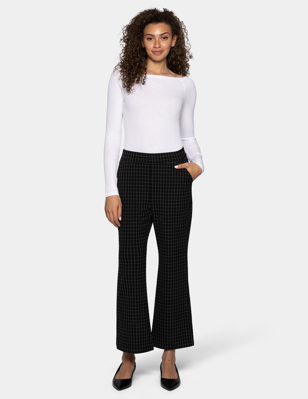 A woman modeling a white off-the-shoulder long-sleeve top and black flare pants with a subtle grid pattern, paired with black flats, against a light gray studio background.