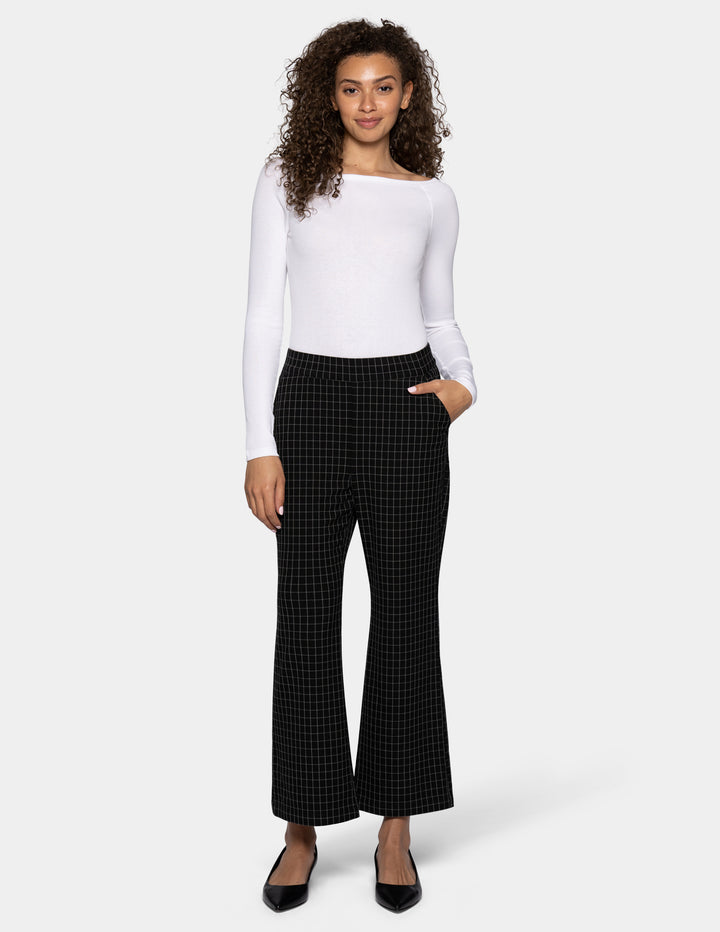 A woman modeling a white off-the-shoulder long-sleeve top and black flare pants with a subtle grid pattern, paired with black flats, against a light gray studio background.