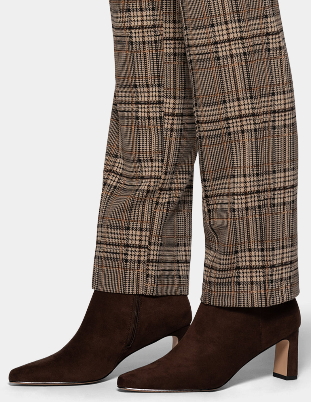 A close-up of a model wearing brown plaid pants styled with suede brown boots, against a plain gray studio background.