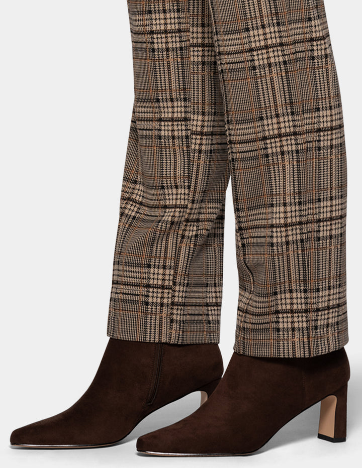 A close-up of a model wearing brown plaid pants styled with suede brown boots, against a plain gray studio background.