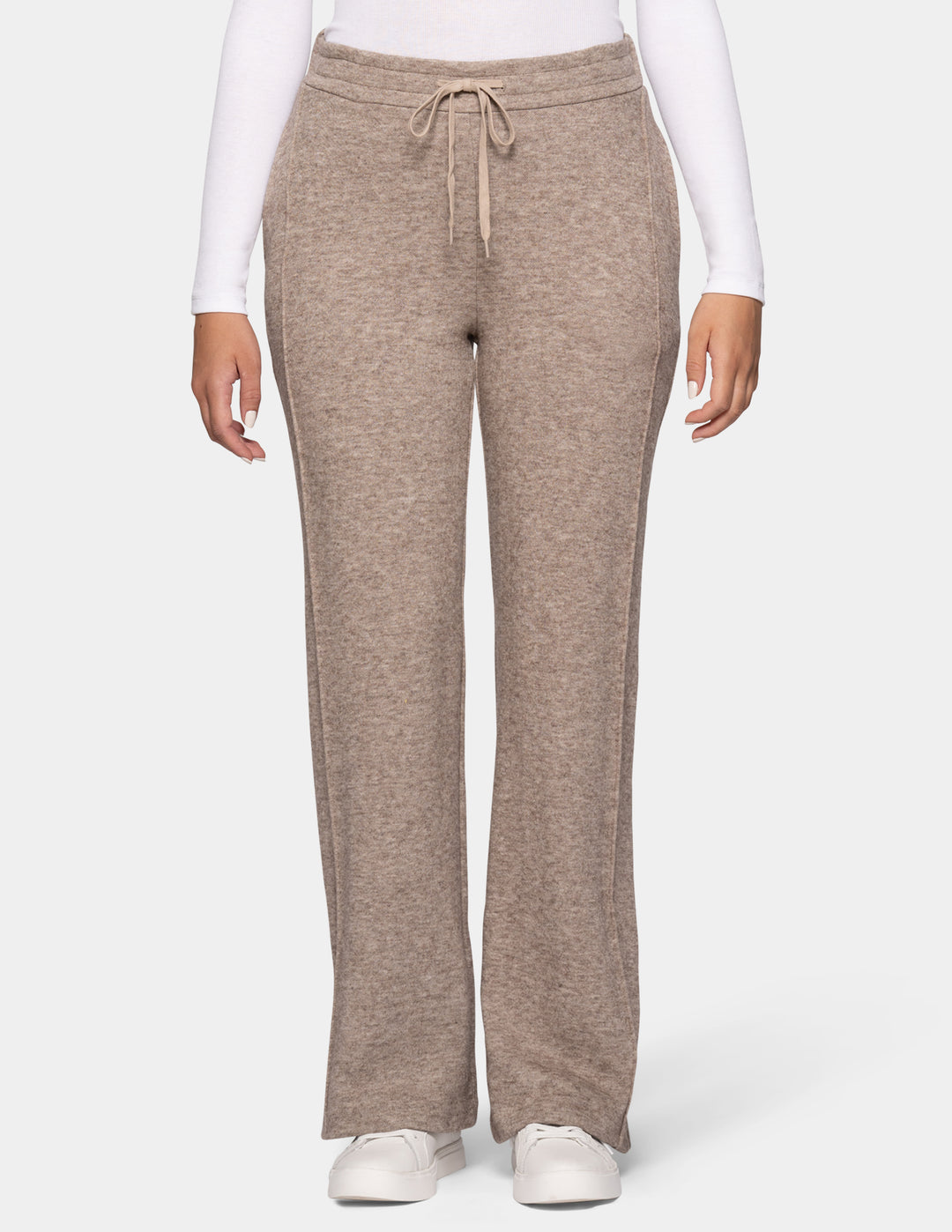 A model wearing beige trackpants and a long sleeve white top, pair with white sneakers against a light grey background.