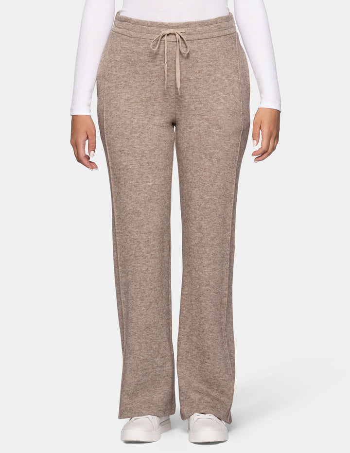 A model wearing beige trackpants and a long sleeve white top, pair with white sneakers against a light grey background.