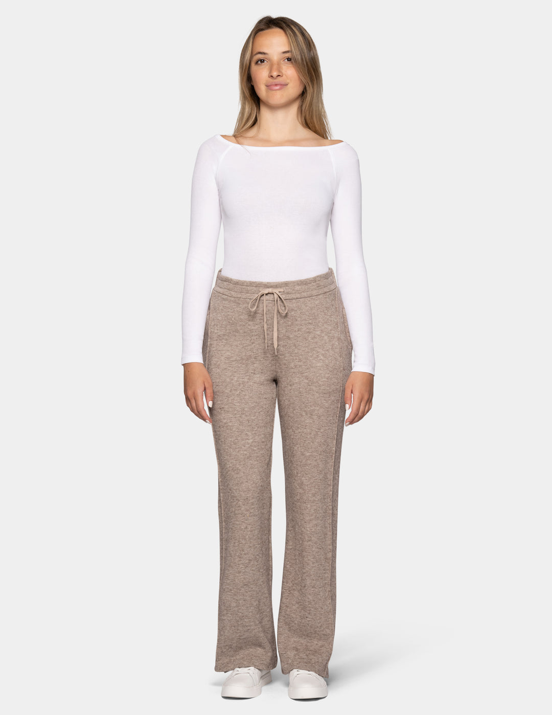 A model wearing beige trackpants and a long sleeve white top, pair with white sneakers against a light grey background.