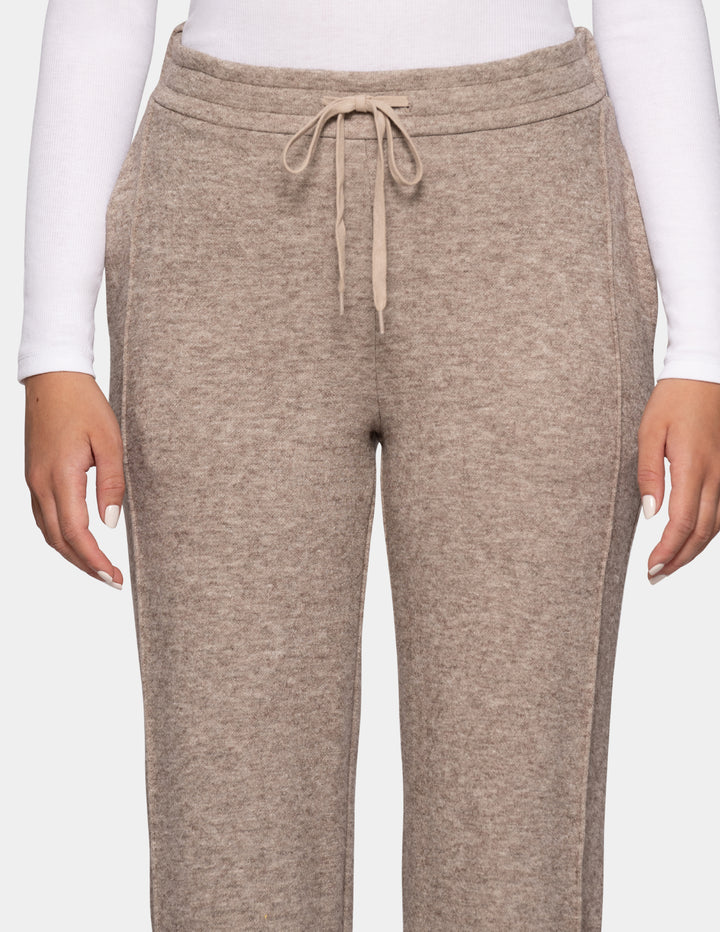 A close-up of a model wearing beige trackpants and a long sleeve white top against a light grey background.